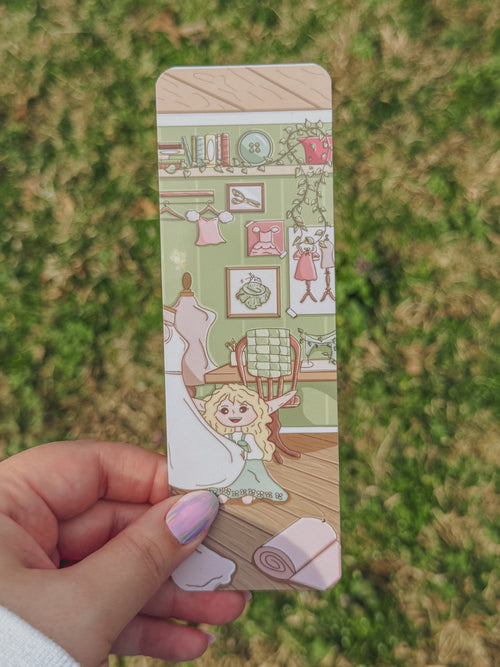 Hand holding Celeste's sewing room bookmark with illustrations on a grassy background