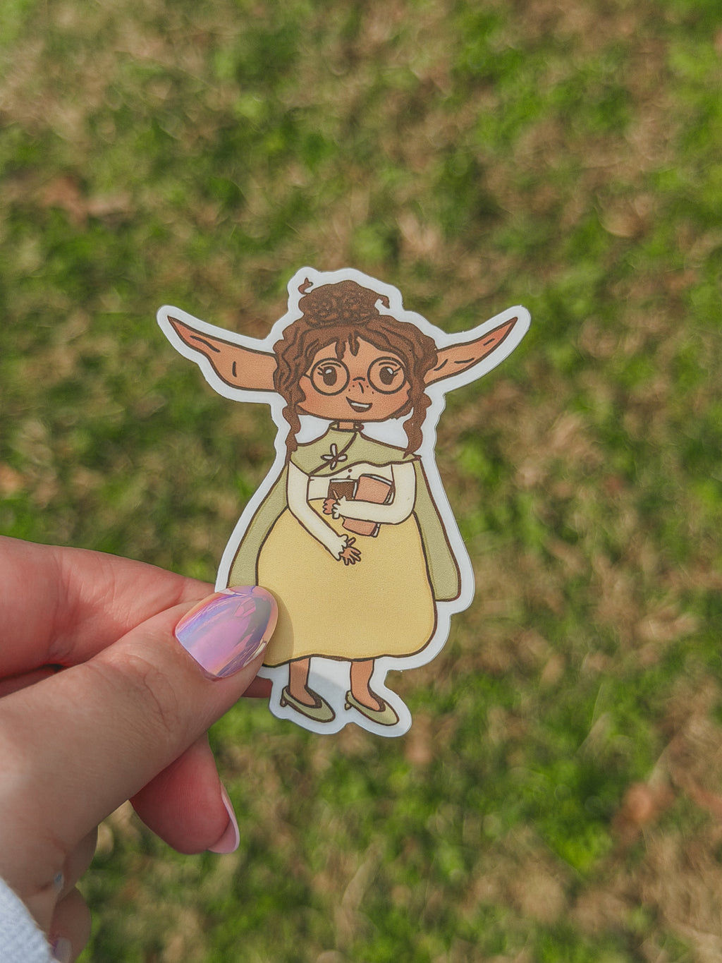 Hand holding a sticker of Junnie with large ears against a grassy background