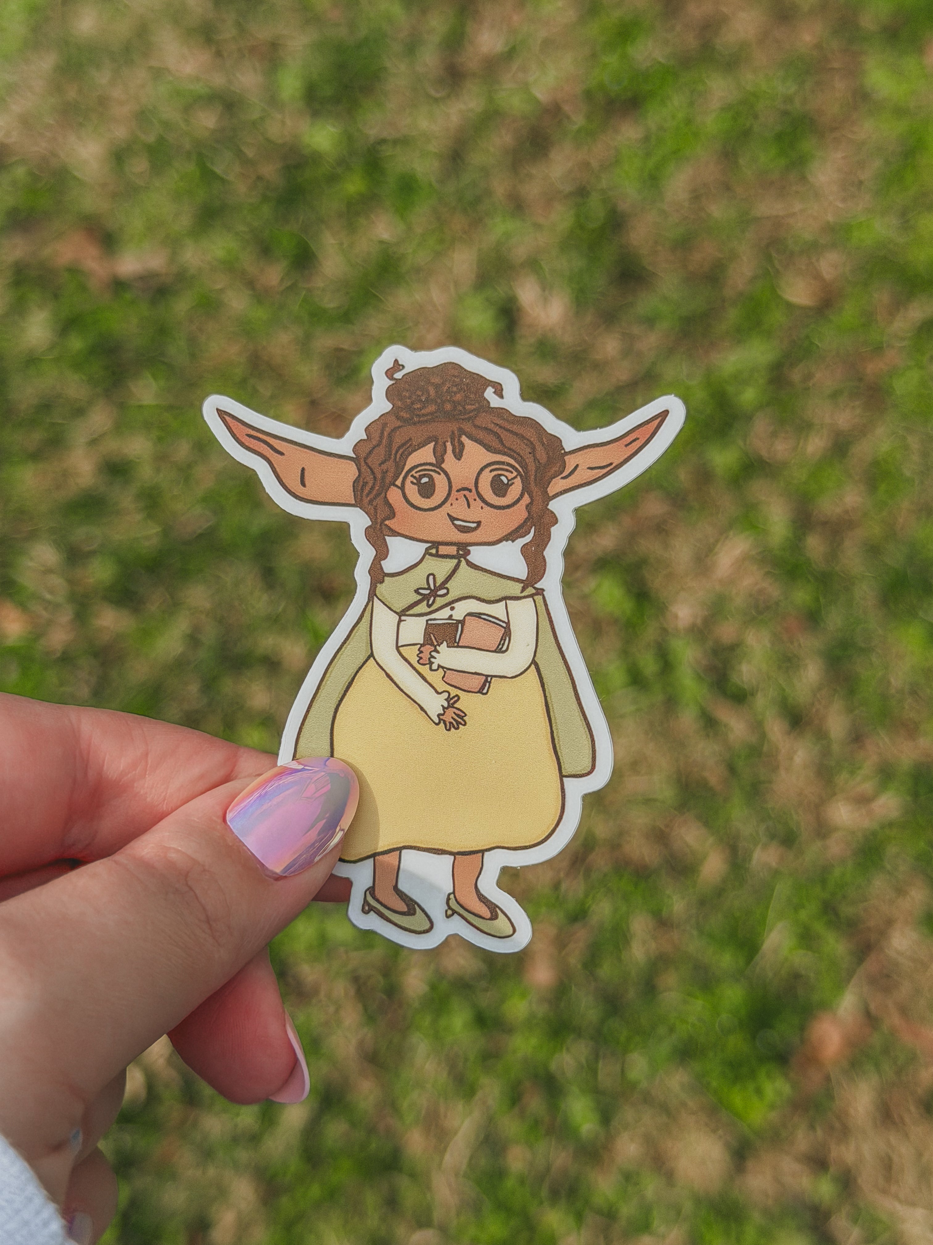 Hand holding a sticker of Junnie with large ears against a grassy background