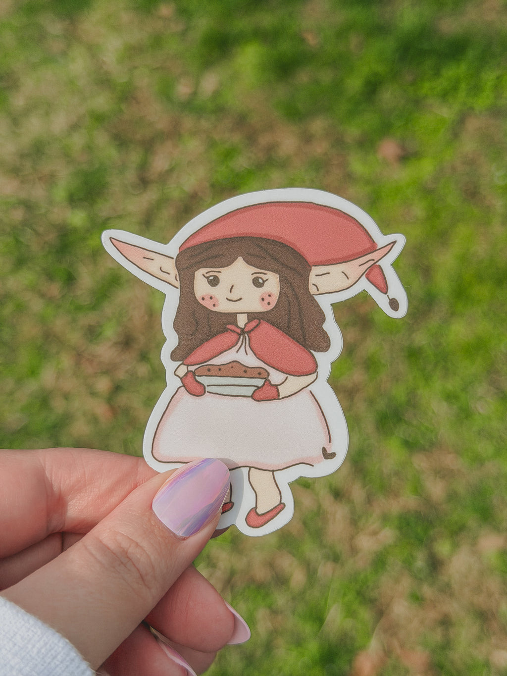 Hand holding a  Meadow sticker with a dark pink hat against a grassy background