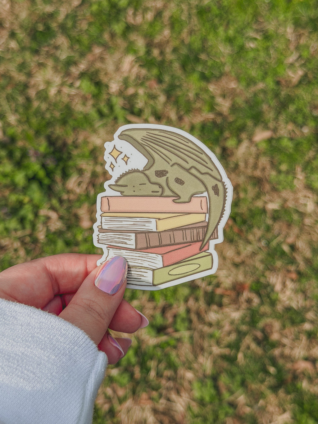 Hand holding a sticker of a dragon with books underneath against a grassy background