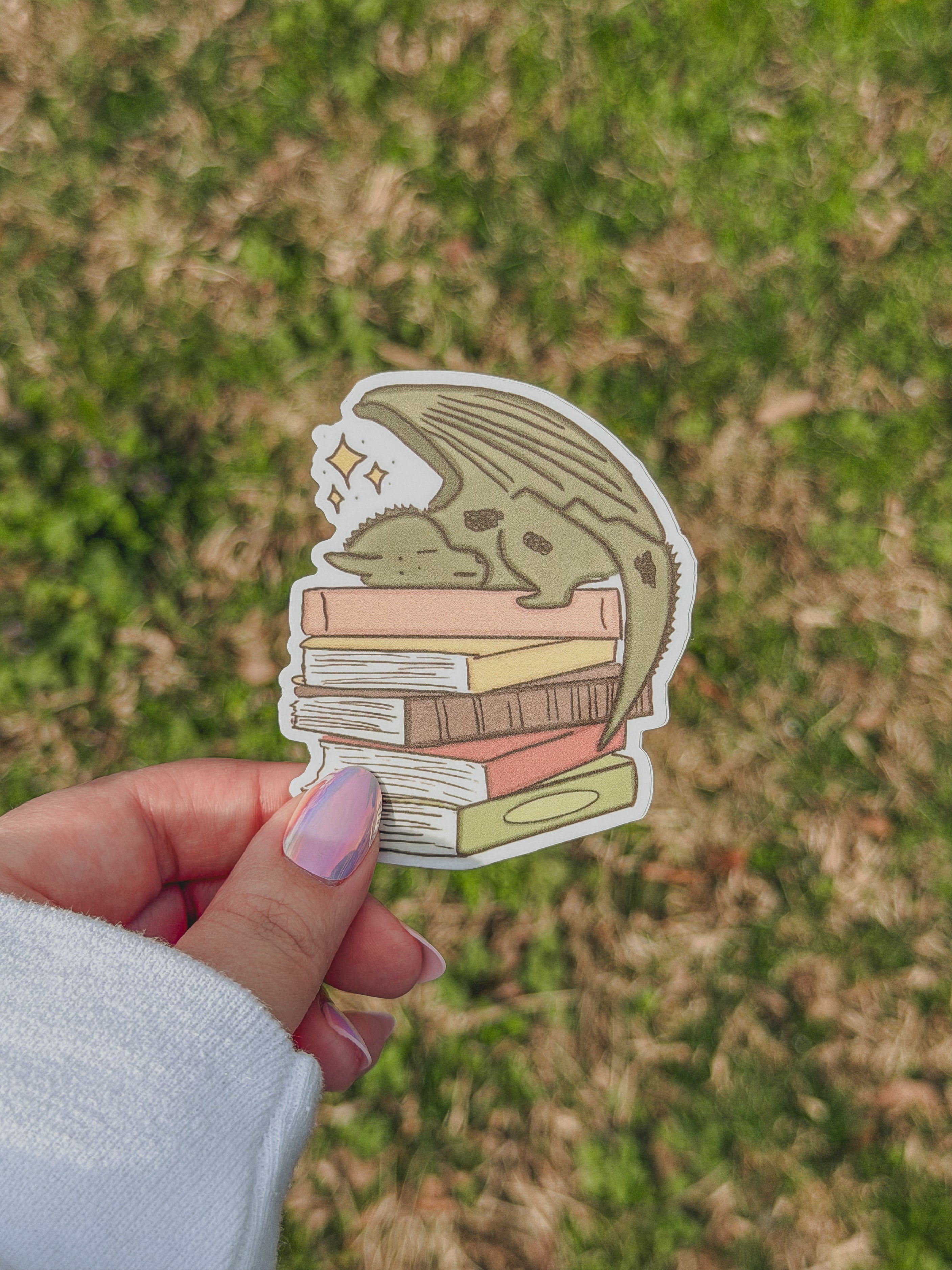 Hand holding a sticker of a dragon with books underneath against a grassy background