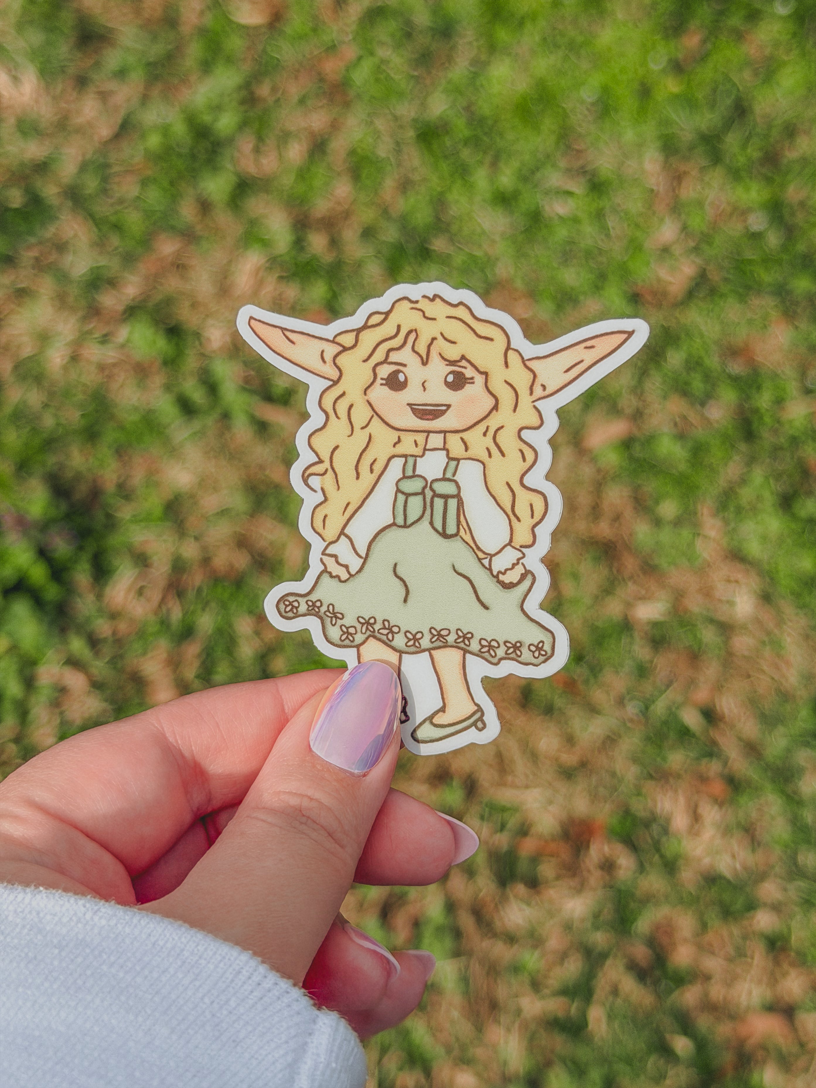 Hand holding a Celeste sticker fairy sticker against a grassy background