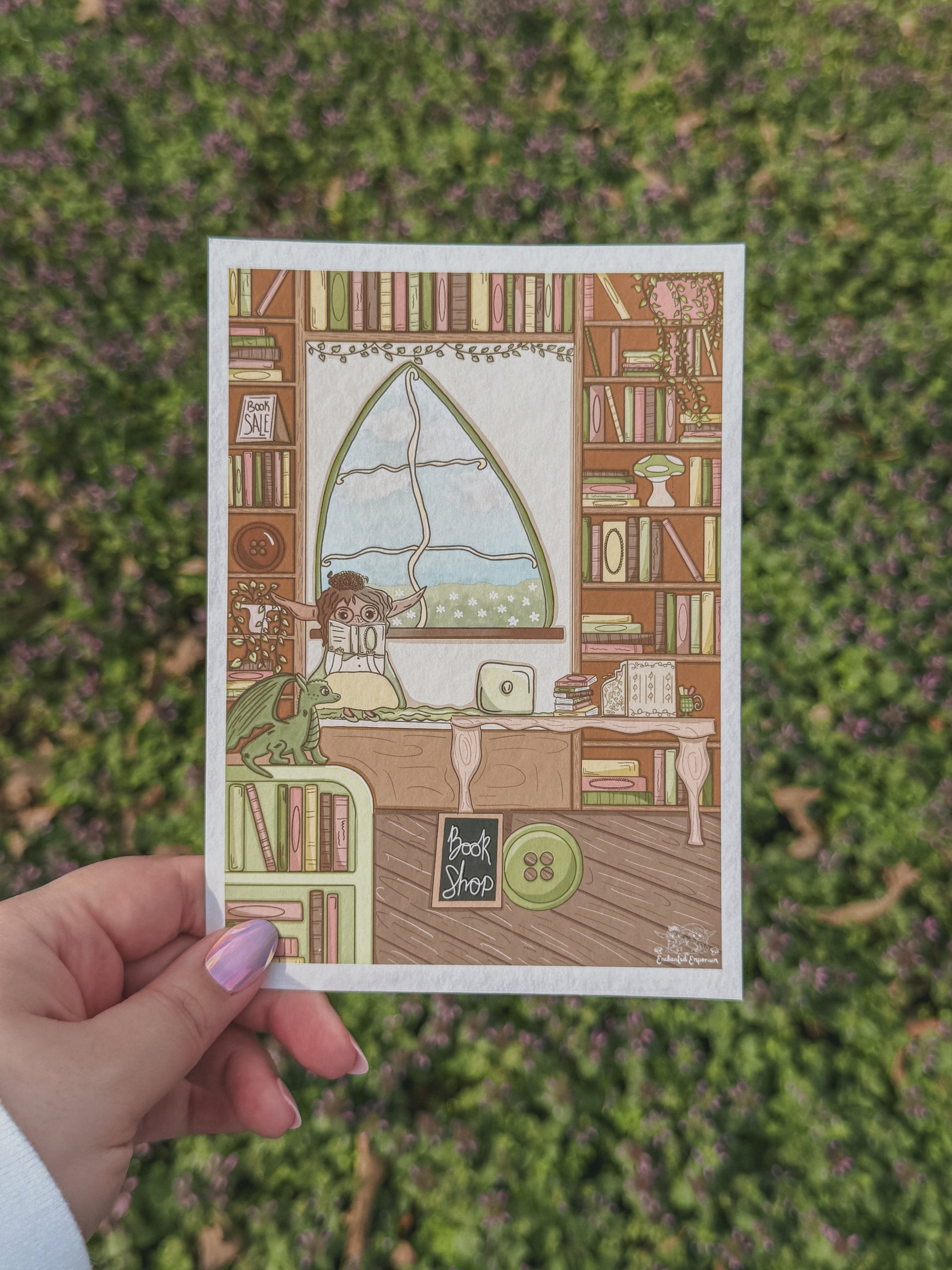 Hand holding a card with a illustration of a bookshop scene against a green leafy background