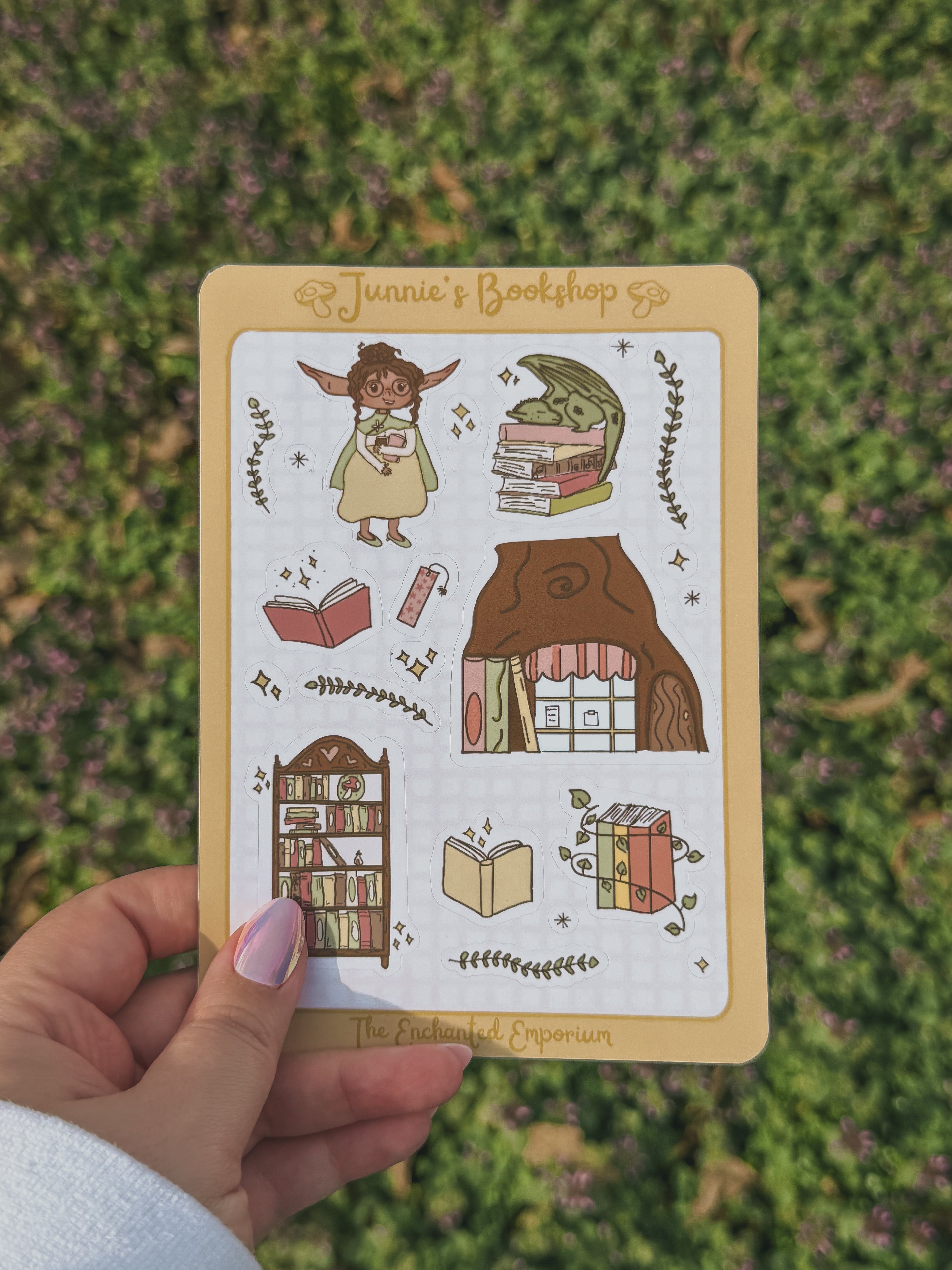 Junnie's sticker sheet illustrations of a bookshop and books held by a hand on a grassy background